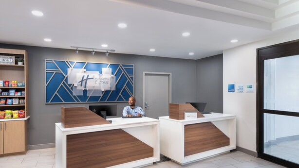 A reception area with two wooden reception desks, a blue geometric wall art, a shelf with snacks, and a door to the right. - Holiday Inn Express Atlanta West - Theme Park Area by IHG (Lithia Springs)