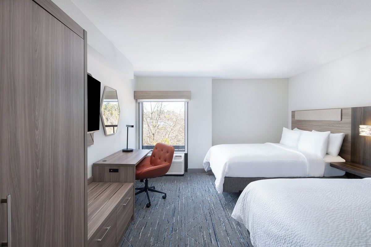 A hotel room with a desk, chair, two beds, a large mirror, and a window with a view of trees.