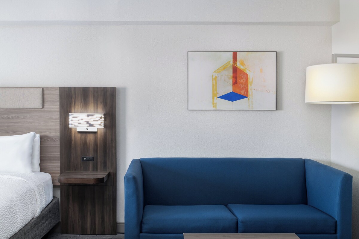 A hotel room with a bed, a blue sofa, a wall-mounted lamp, and a painting.