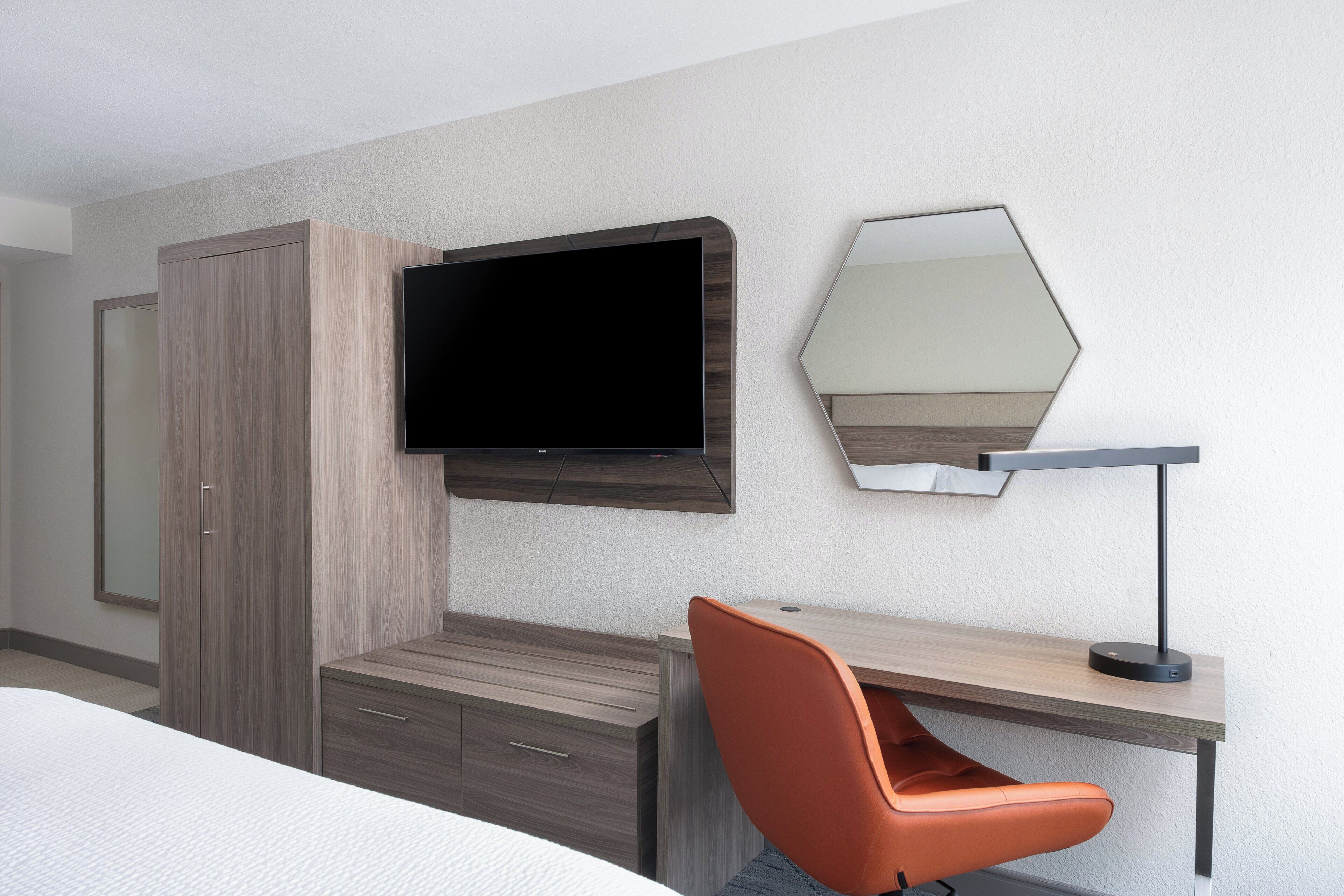 Habitación de hotel con una cama, un escritorio con silla, televisión, espejo y armario.
