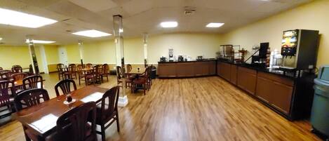 A dining area with wooden tables and chairs, a kitchen counter with appliances, and a high ceiling with fluorescent lighting.