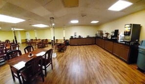 A dining area with wooden tables and chairs, a kitchen counter with appliances, and a high ceiling with fluorescent lighting.