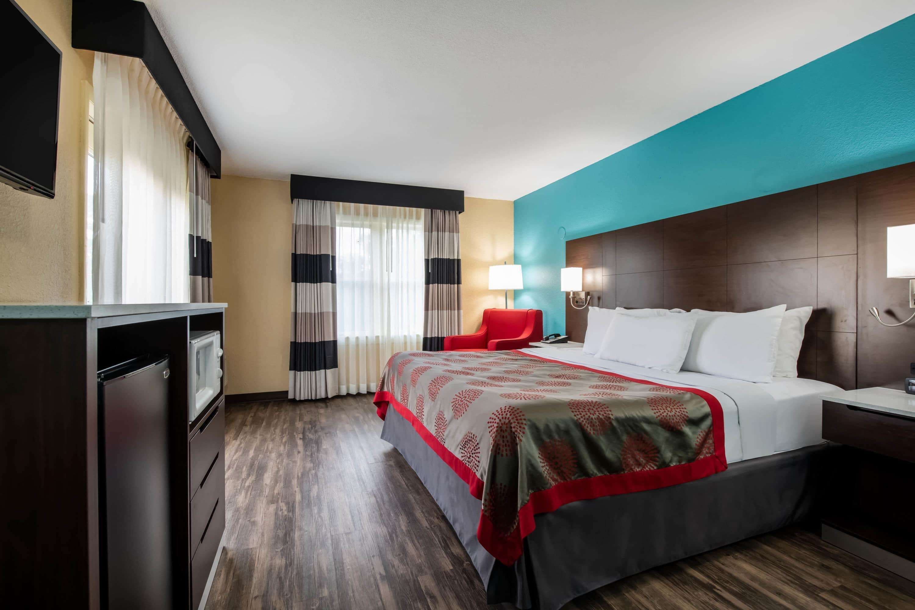 A hotel room with a bed, a red chair, a desk, a lamp, and a TV.