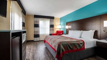 A hotel room with a bed, a red chair, a desk, a lamp, and a TV.