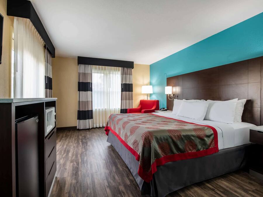 A hotel room with a bed, a red chair, a desk, a lamp, and a TV.