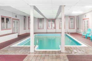An indoor swimming pool with clear water, surrounded by white walls with red trim, and two turquoise chairs near the pool.