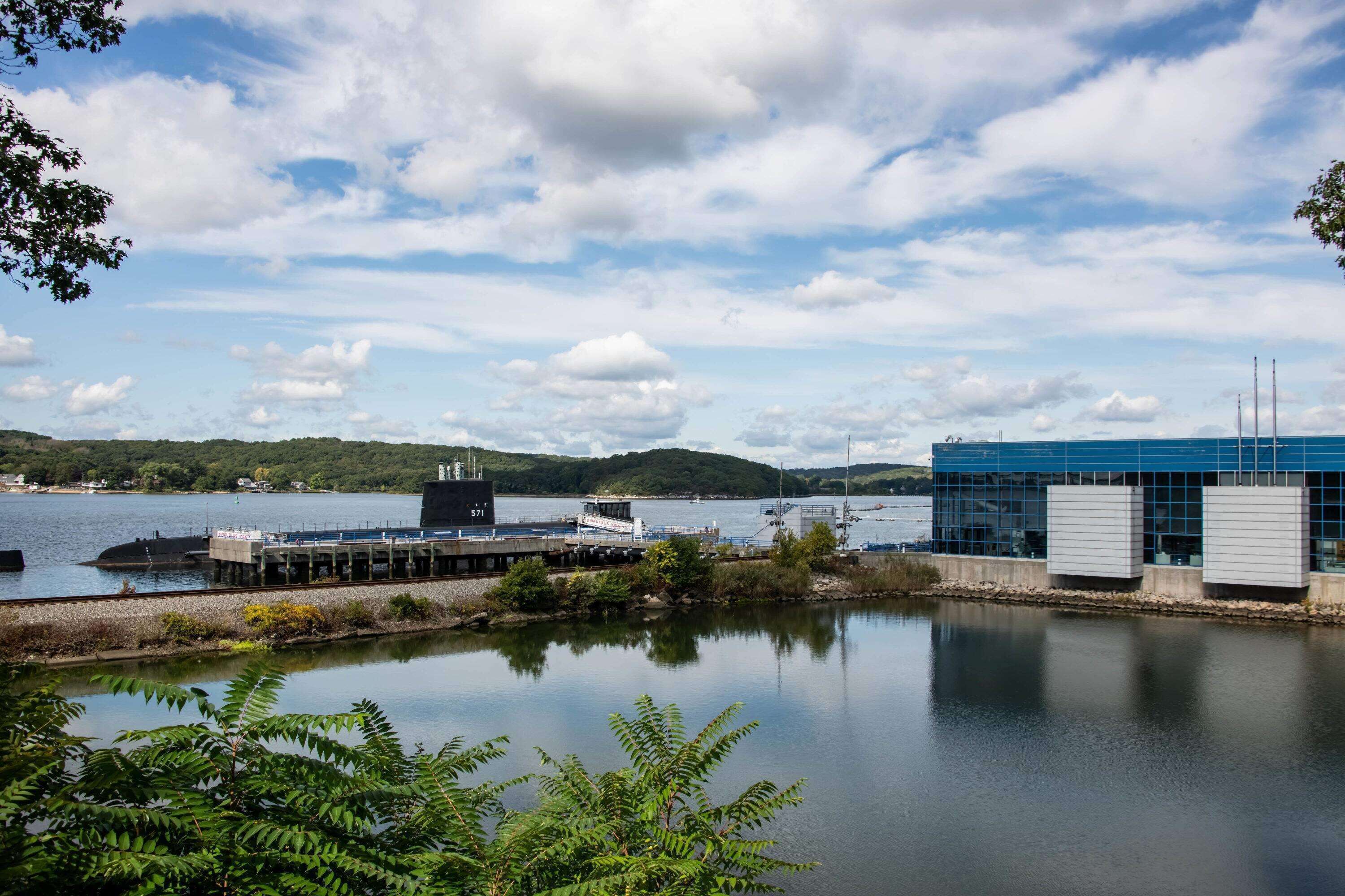 A waterfront industrial facility with a large building, dock, and calm water reflecting the sky.