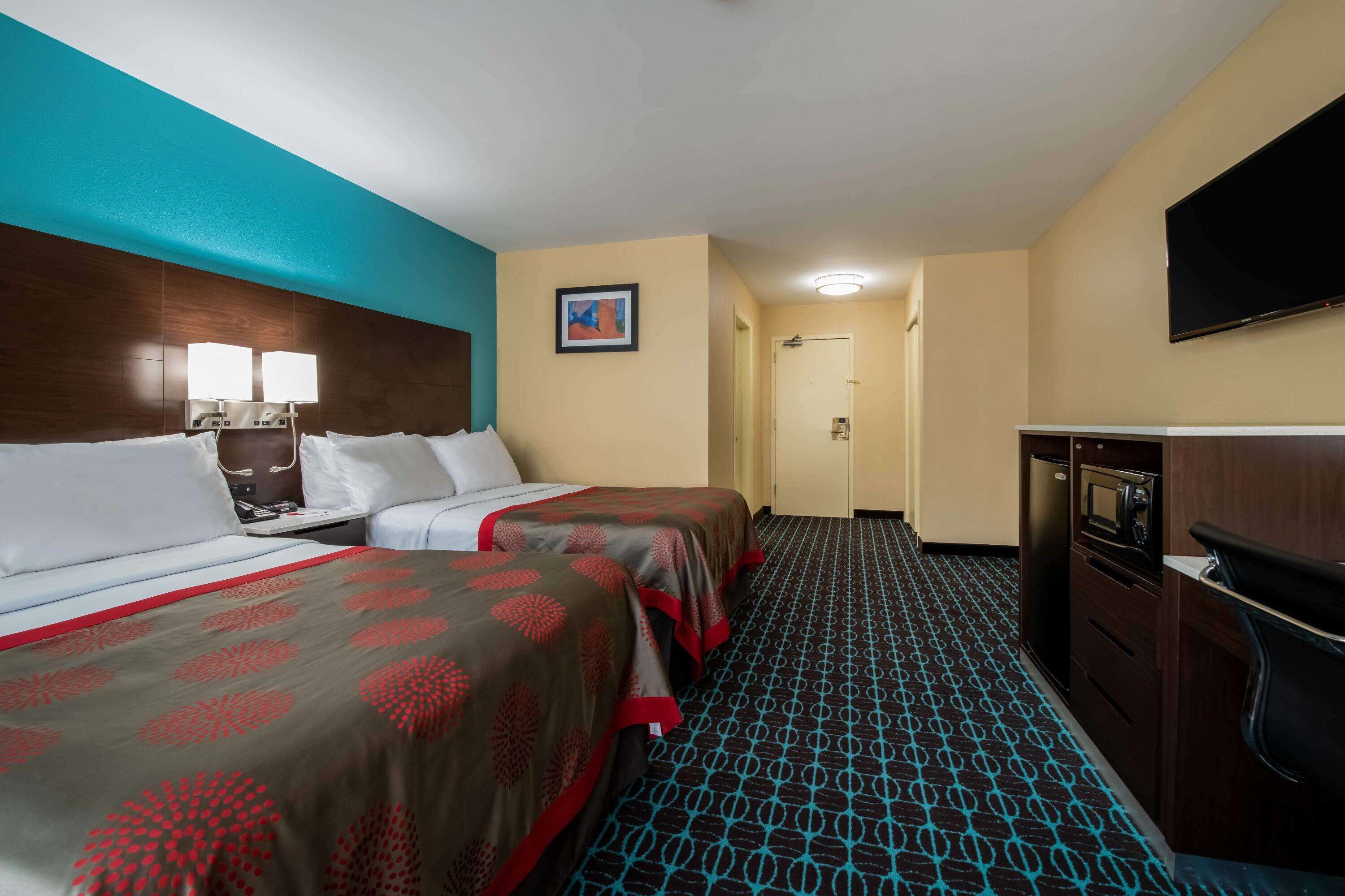 A hotel room with two beds, a desk with a lamp, a flat-screen TV, a microwave, and a door leading to a hallway.