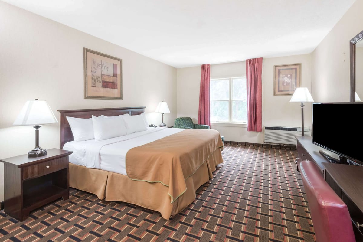A hotel room with a bed, two bedside tables with lamps, a television, a window with curtains, and a patterned carpet.