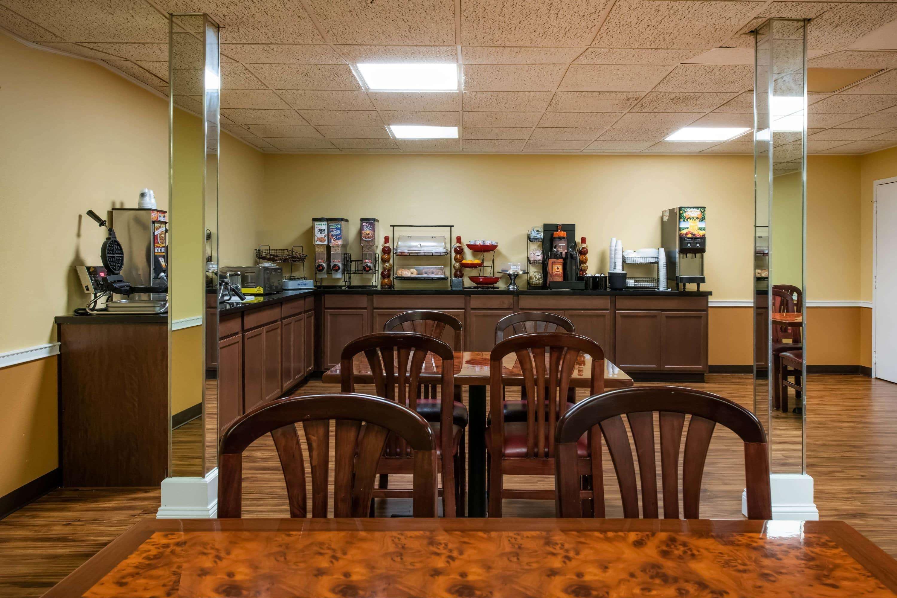 A dining area with a wooden table and chairs, a counter with various food and beverage dispensers, and a kitchen area in the background.