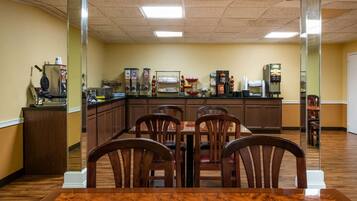 A dining area with a wooden table and chairs, a counter with various food and beverage dispensers, and a kitchen area in the background.
