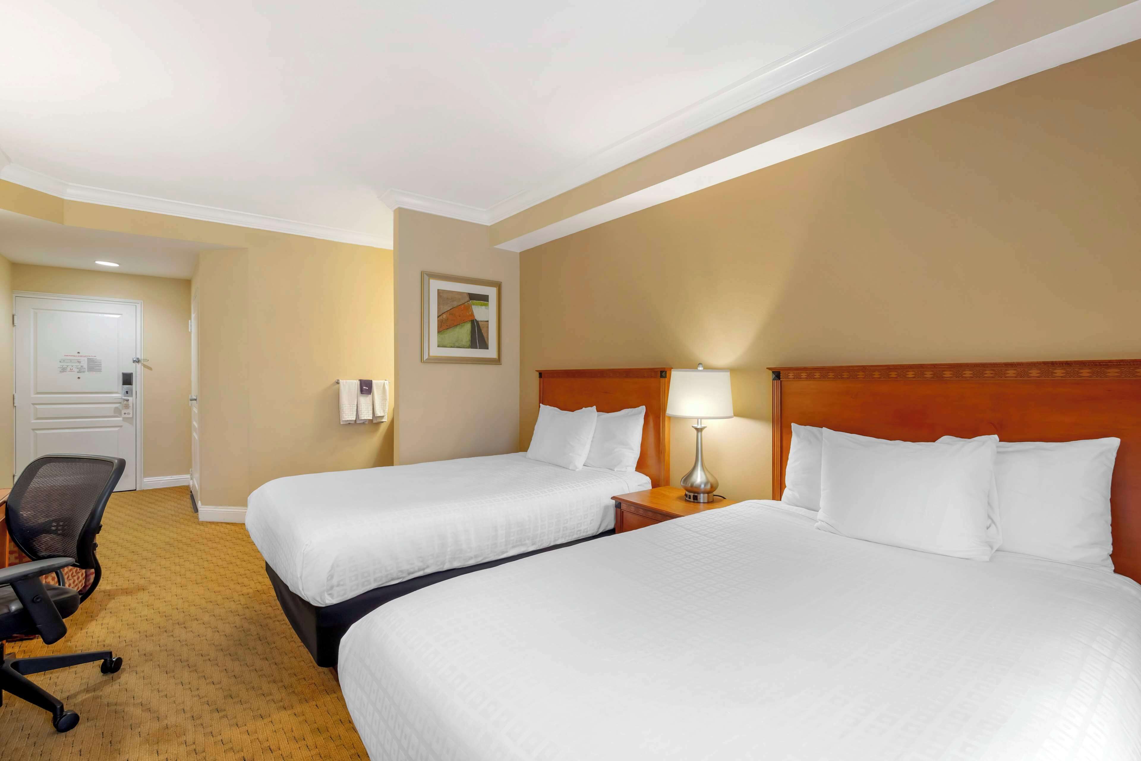 A hotel room with two beds, a desk, a chair, and a lamp.