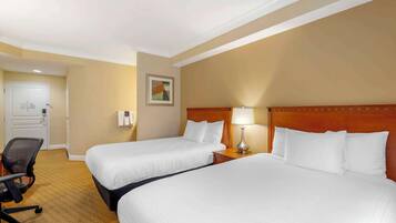 A hotel room with two beds, a desk, a chair, and a lamp.