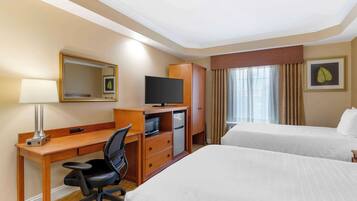 A hotel room with a wooden desk, a television, a microwave, a refrigerator, two beds, a chair, a lamp, and a window with curtains.