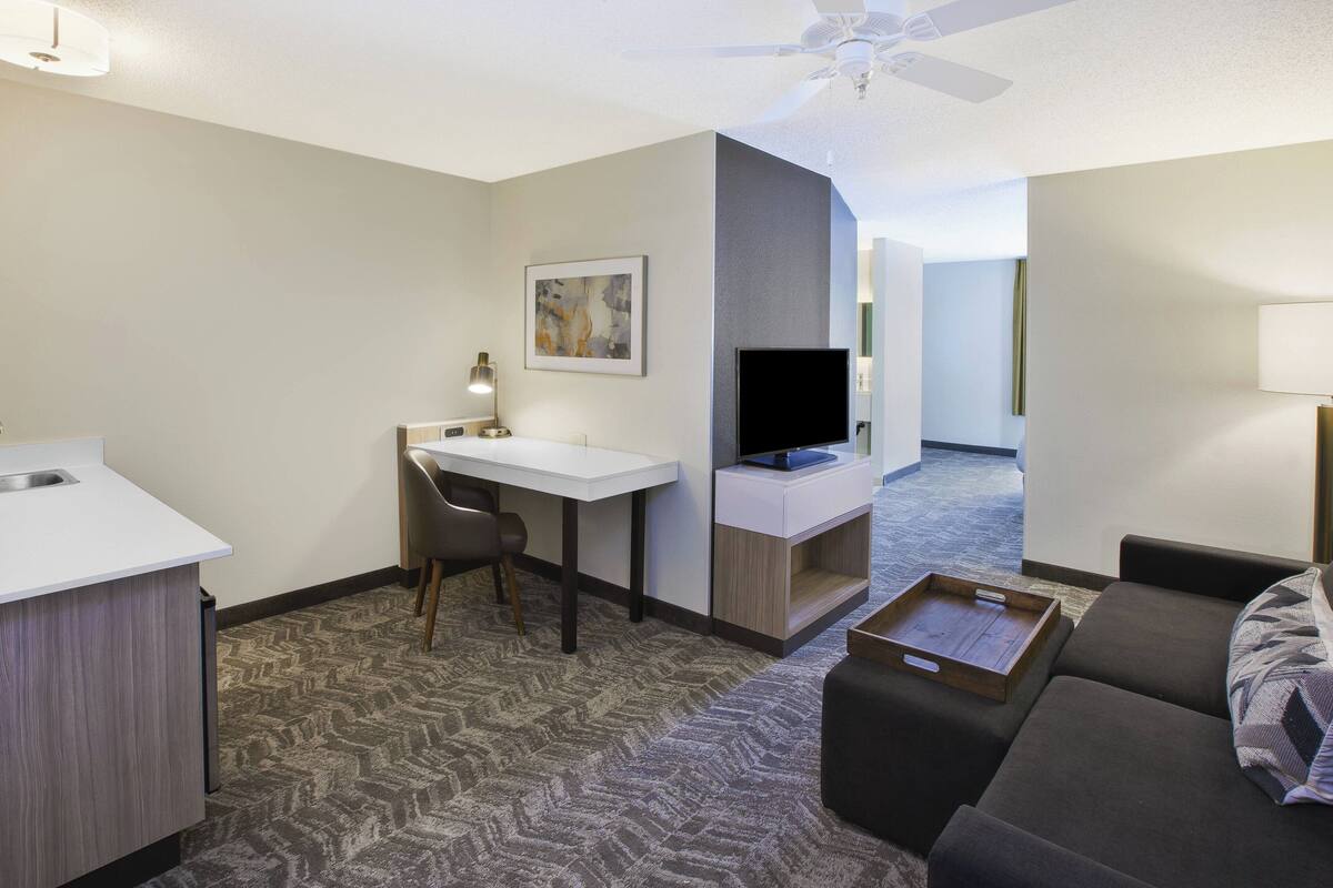 A compact hotel room with a kitchenette, a desk, a flat-screen TV, a sofa, and a ceiling fan.