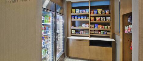 Una pequeña kitchenette con refrigerador, microondas y un estante repleto de botanas y bebidas.