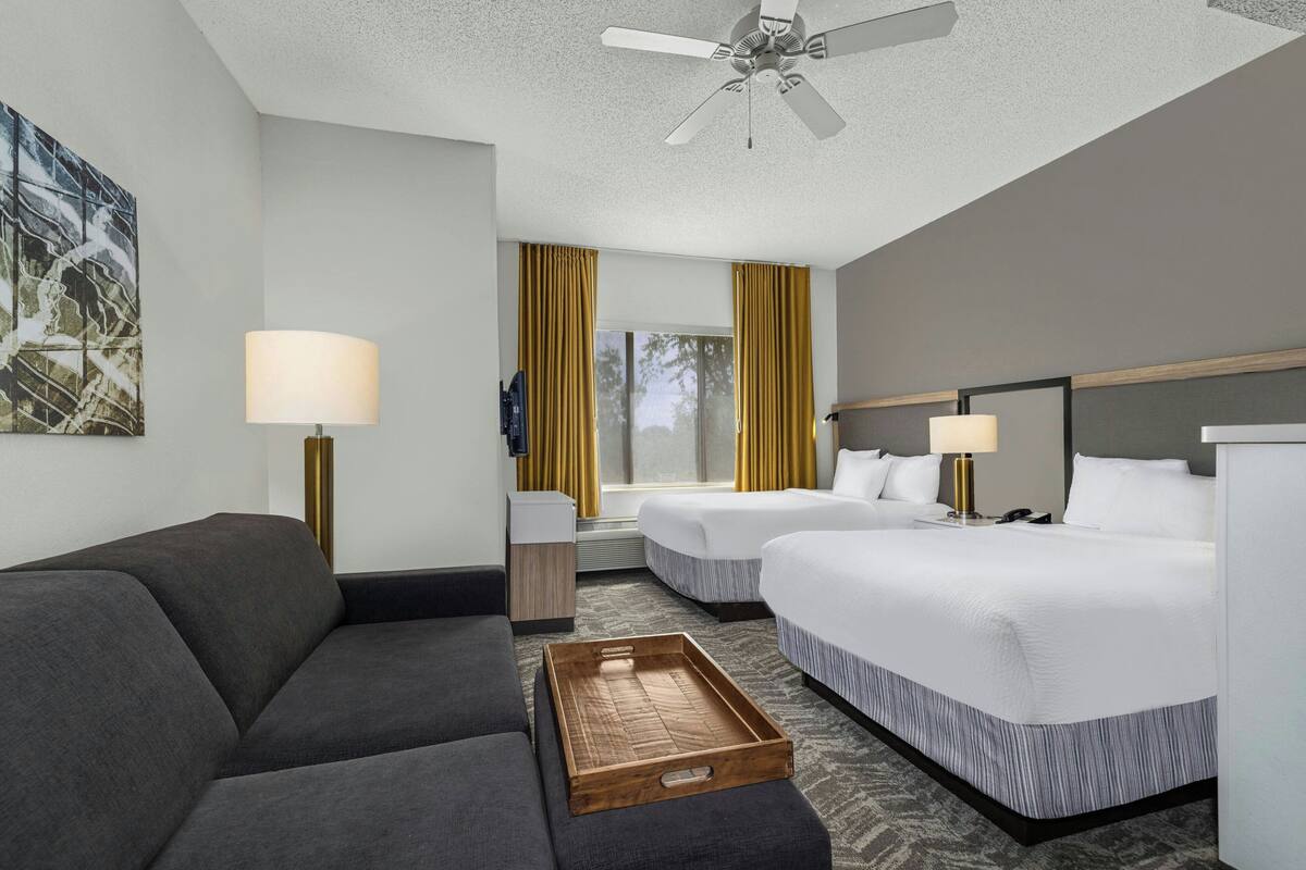 A hotel room with a sofa, two beds, a ceiling fan, a lamp, and a tray table.
