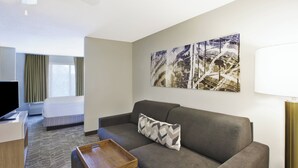 Una habitación de hotel moderna con un sofá gris, una mesita de madera, una cama con edredón blanco y un cuadro abstracto grande en la pared.