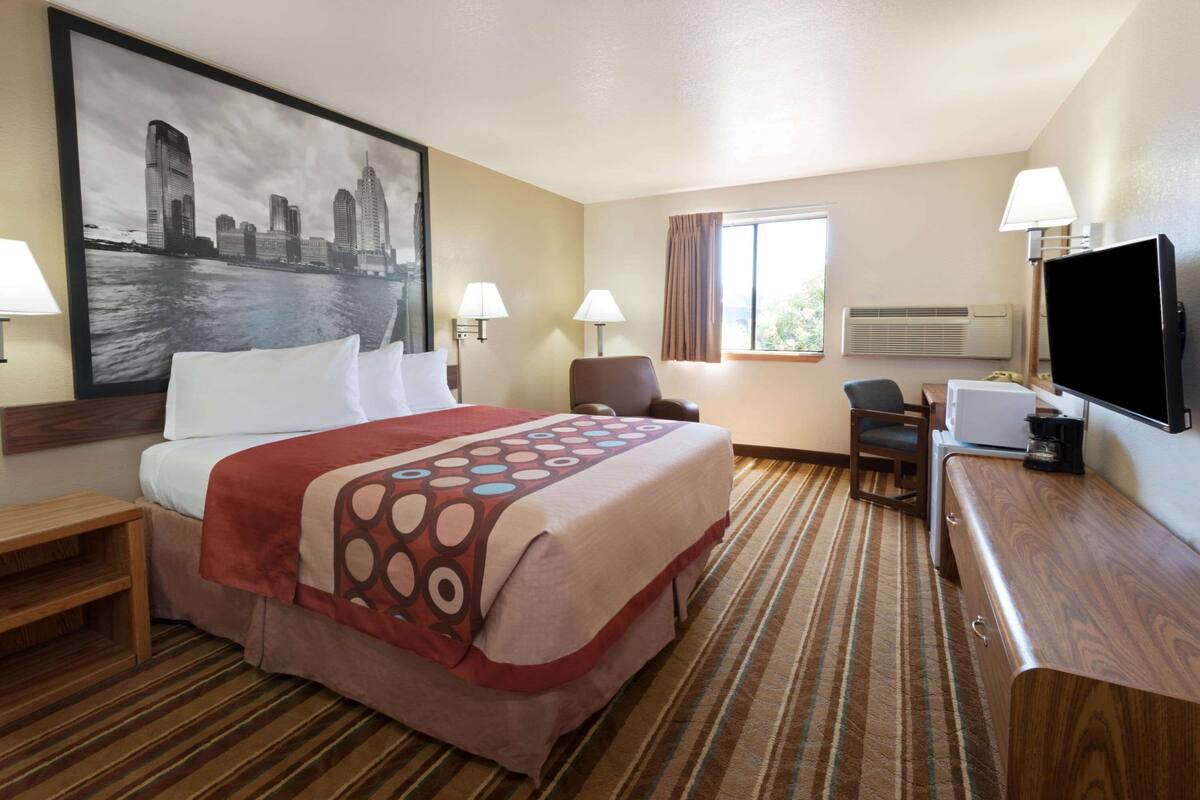 Habitación de hotel con cama, dos sillas, un escritorio, un televisor y un cuadro grande con una vista urbana.