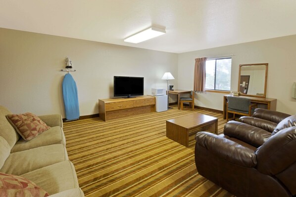 A hotel room with a sofa, a brown leather chair, a television, a desk with a lamp, a microwave, and a window with curtains.