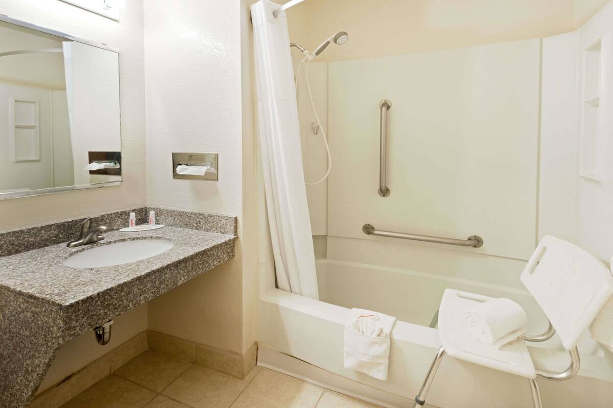 Un baño con lavamanos de granito, espejo, ducha con barra de apoyo y bañera blanca.