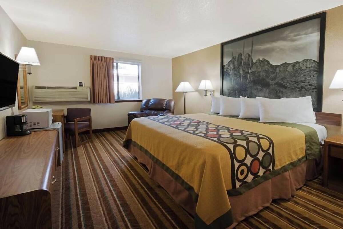 Habitación de hotel con cama, escritorio con lámpara, silla, televisor y un cuadro en la pared.