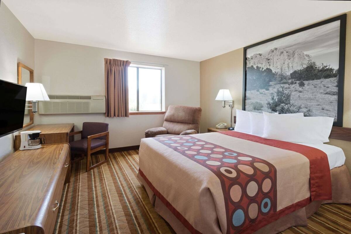 Habitación de hotel con cama, escritorio con cafetera, silla, televisor y ventana con cortinas.