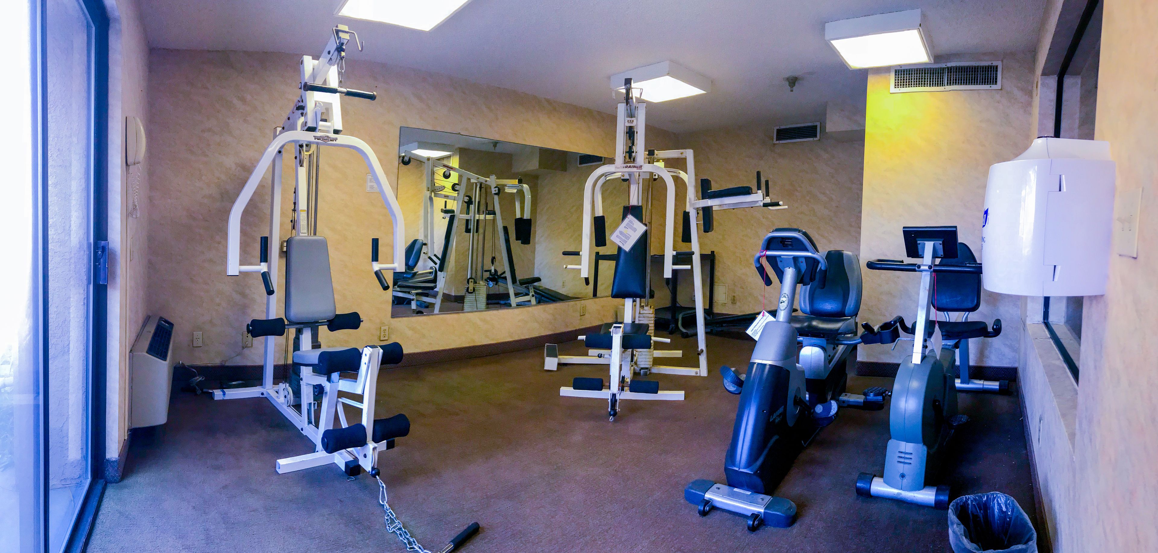 Un gimnasio con diferentes equipos de ejercicio, un espejo grande y un aire acondicionado instalado en la pared.
