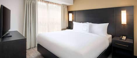 Habitación de hotel con una cama grande, un televisor de pantalla plana, una mesita de noche con lámpara y una ventana con cortinas.