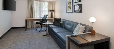 Une chambre d’hôtel moderne avec un canapé gris, une petite table ronde, un bureau avec une chaise, un téléviseur fixé au mur et des œuvres d’art encadrées accrochées au mur.