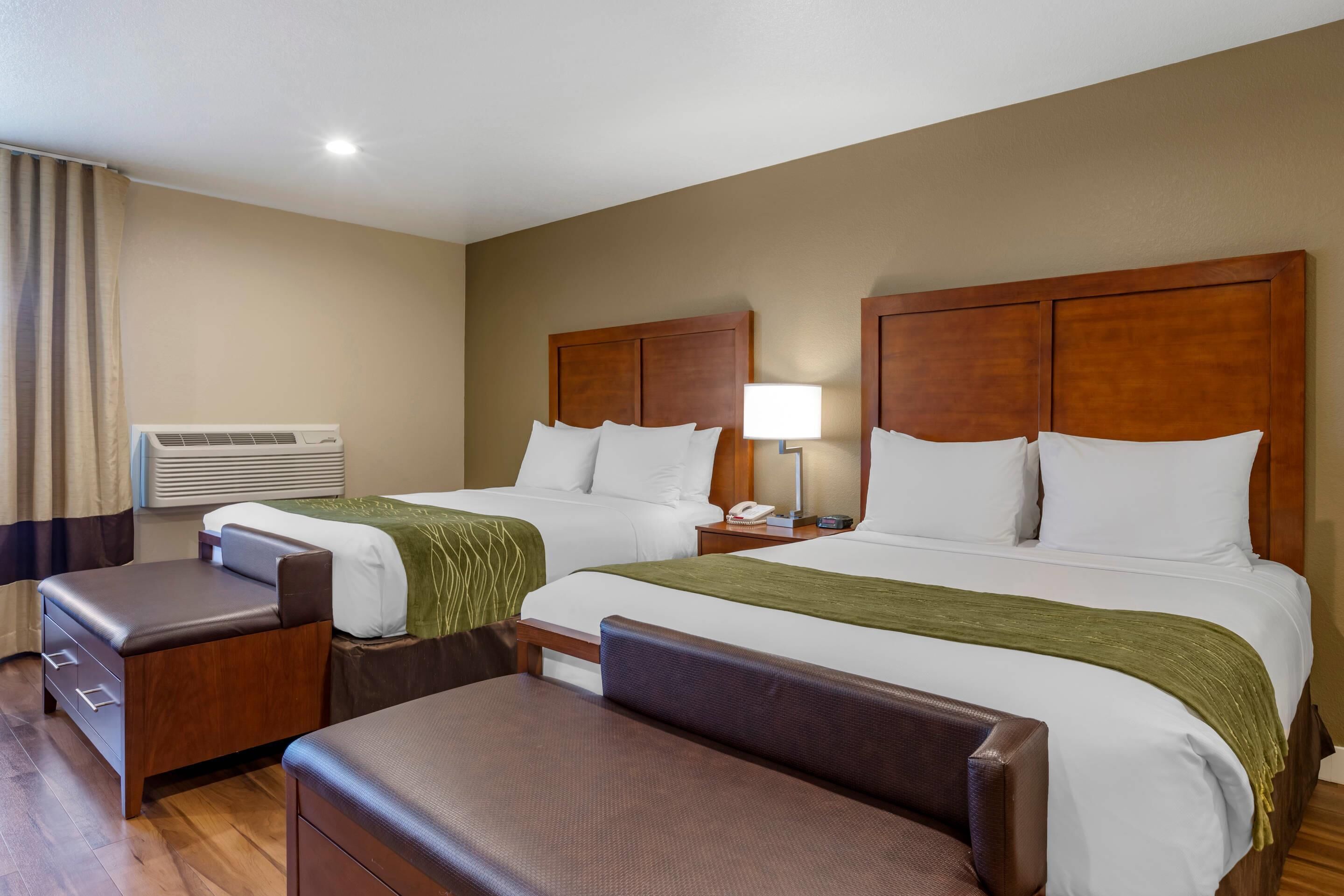 A hotel room with two beds, a desk, a chair, a lamp, and an air conditioning unit.