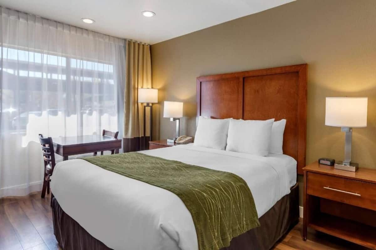 A hotel room with a large bed, two bedside lamps, a desk with a chair, and a window with curtains.