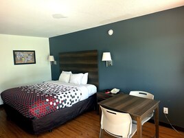 Habitación de hotel con cama, mesita de noche, escritorio, silla, lámpara y teléfono en la pared.