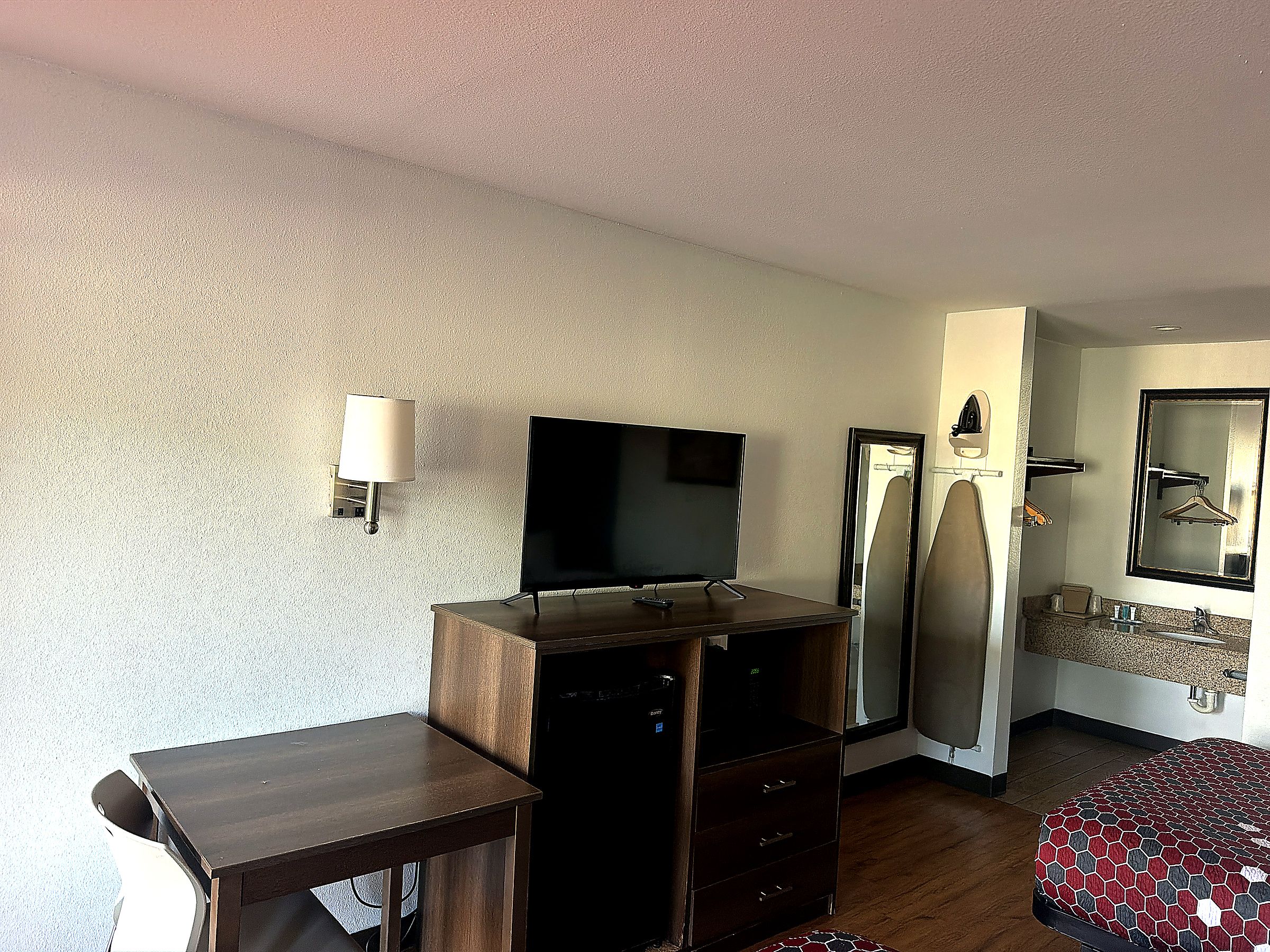 Habitación de hotel con televisor de pantalla plana, cómoda de madera, escritorio, cama con colcha estampada, espejo y lámpara de pared.