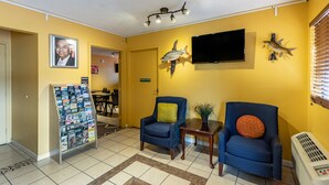 Un área de recepción con dos sillones azules, una pequeña mesa con una planta, un adorno de pez, un televisor montado y un atril para revistas.