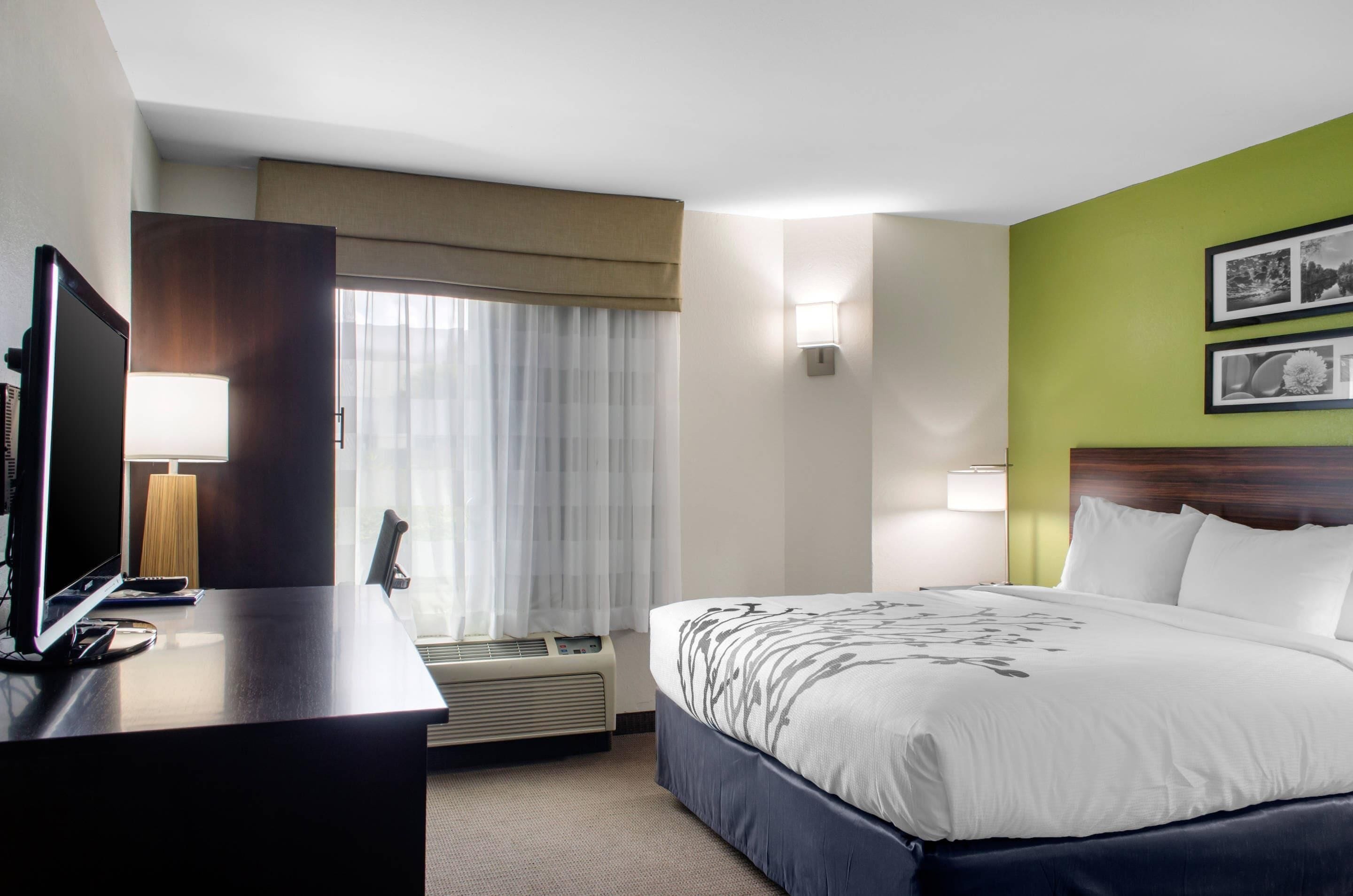 Quarto de hotel com cama, escrivaninha com computador, televisão, janela com cortinas e luminária fixada na parede.