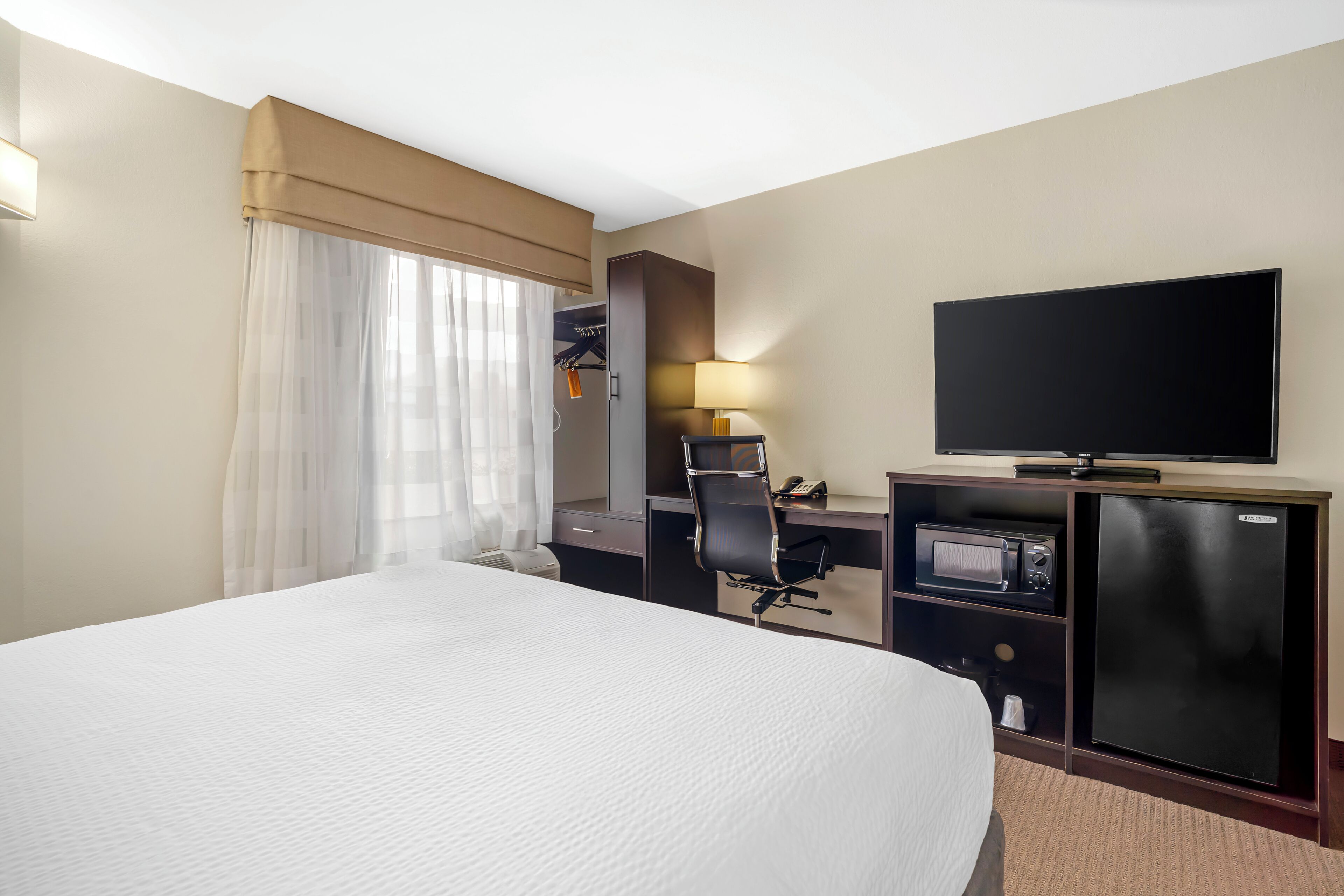 Ein Hotelzimmer mit einem großen Bett, einem Schreibtisch mit Computer, einem Fernseher und einem kleinen Kühlschrank.