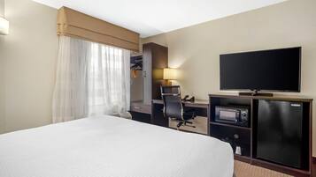 Ein Hotelzimmer mit einem großen Bett, einem Schreibtisch mit Computer, einem Fernseher und einem kleinen Kühlschrank.