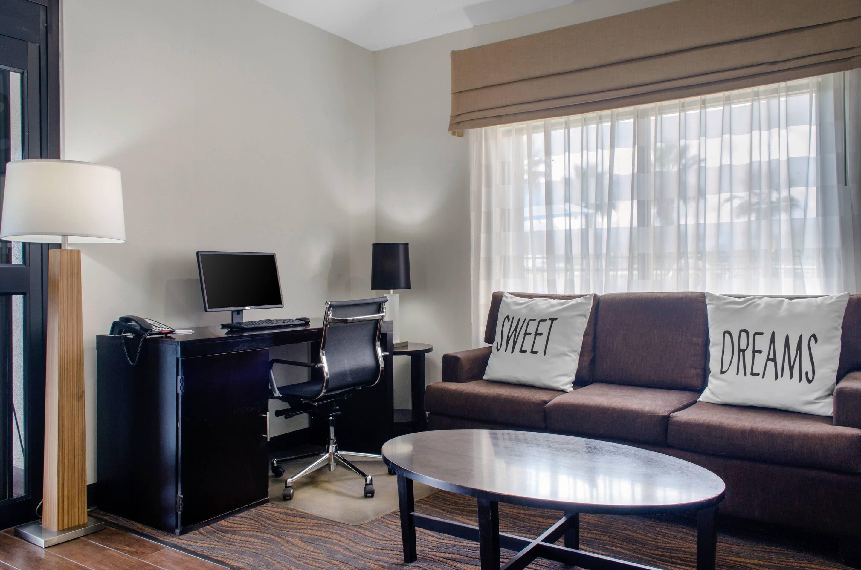 A hotel room with a desk, computer, chair, sofa, and a coffee table.