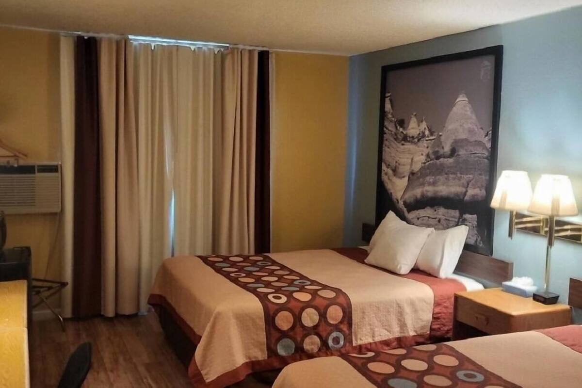 Habitación de hotel con dos camas, un escritorio, una silla, un ventanal con cortinas, una lámpara de pared y un cuadro.