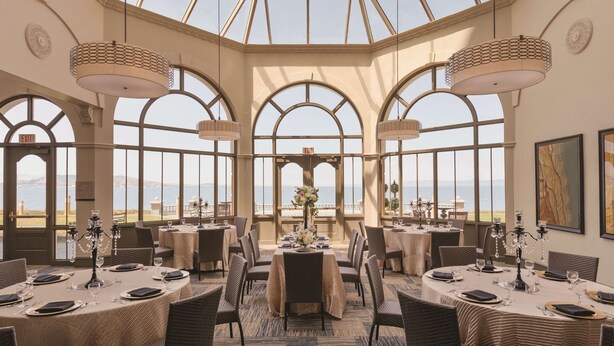A dining area with tables set for service, large arched windows, and a view of the sea.