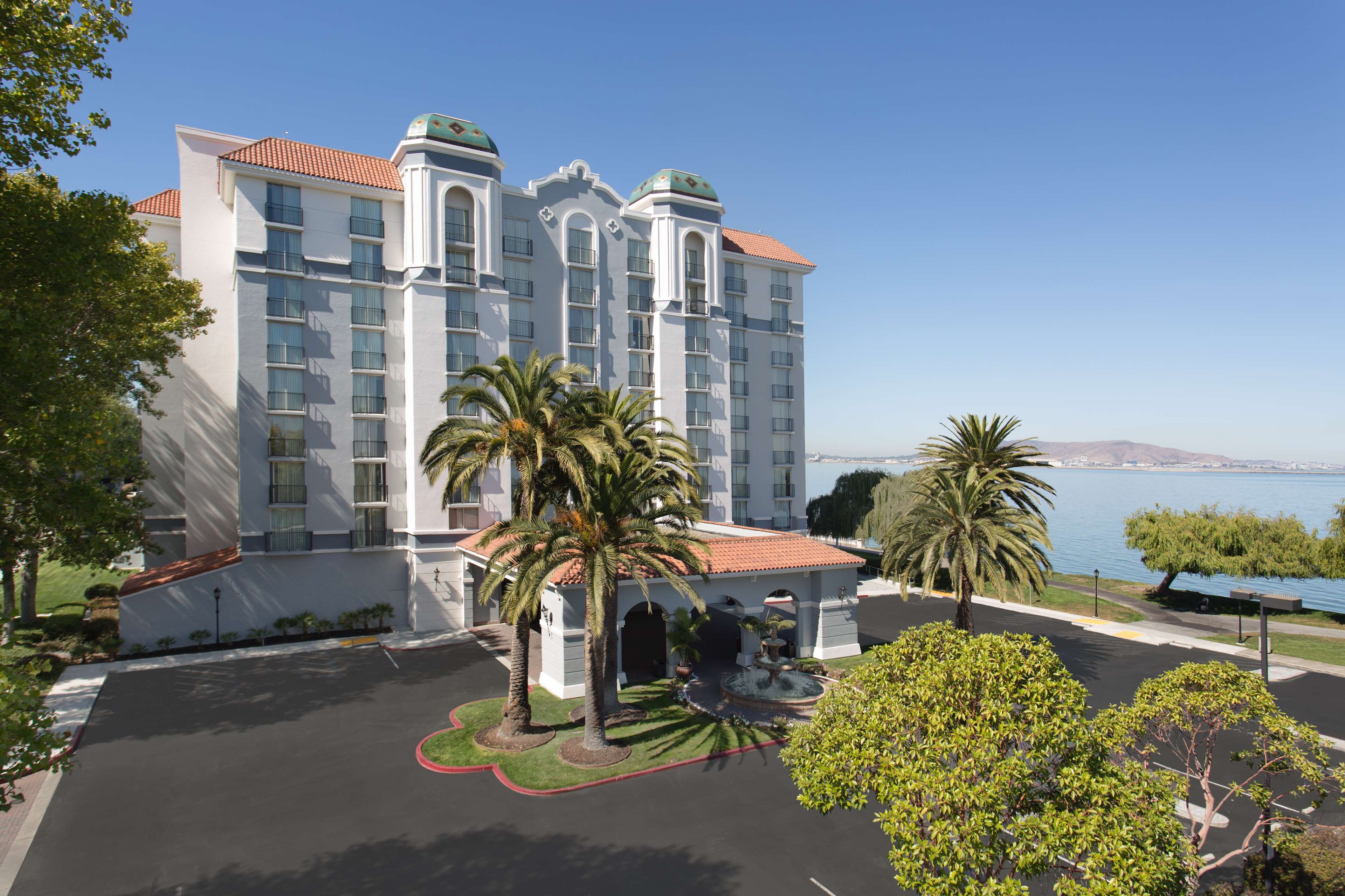 A large hotel with multiple floors, a courtyard with palm trees, and a view of the water.