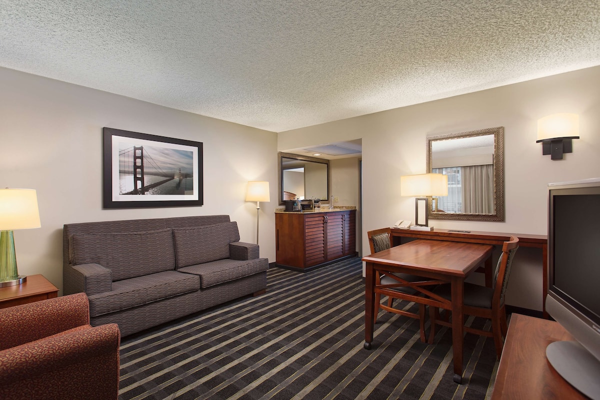 A hotel room with a sofa, a dining table, a TV, a mirror, and a framed picture of a bridge.