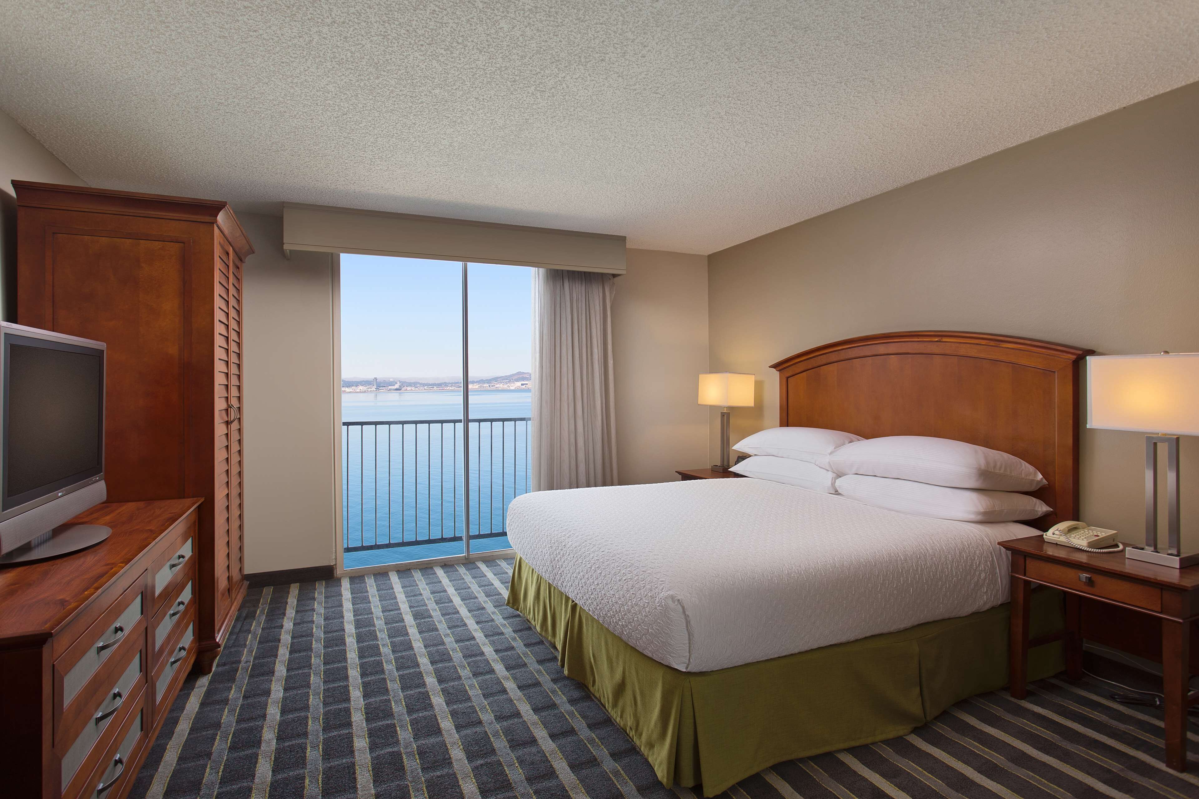 A hotel room with a large bed, a TV on a wooden dresser, a bedside lamp, and a view of the sea through a large window.