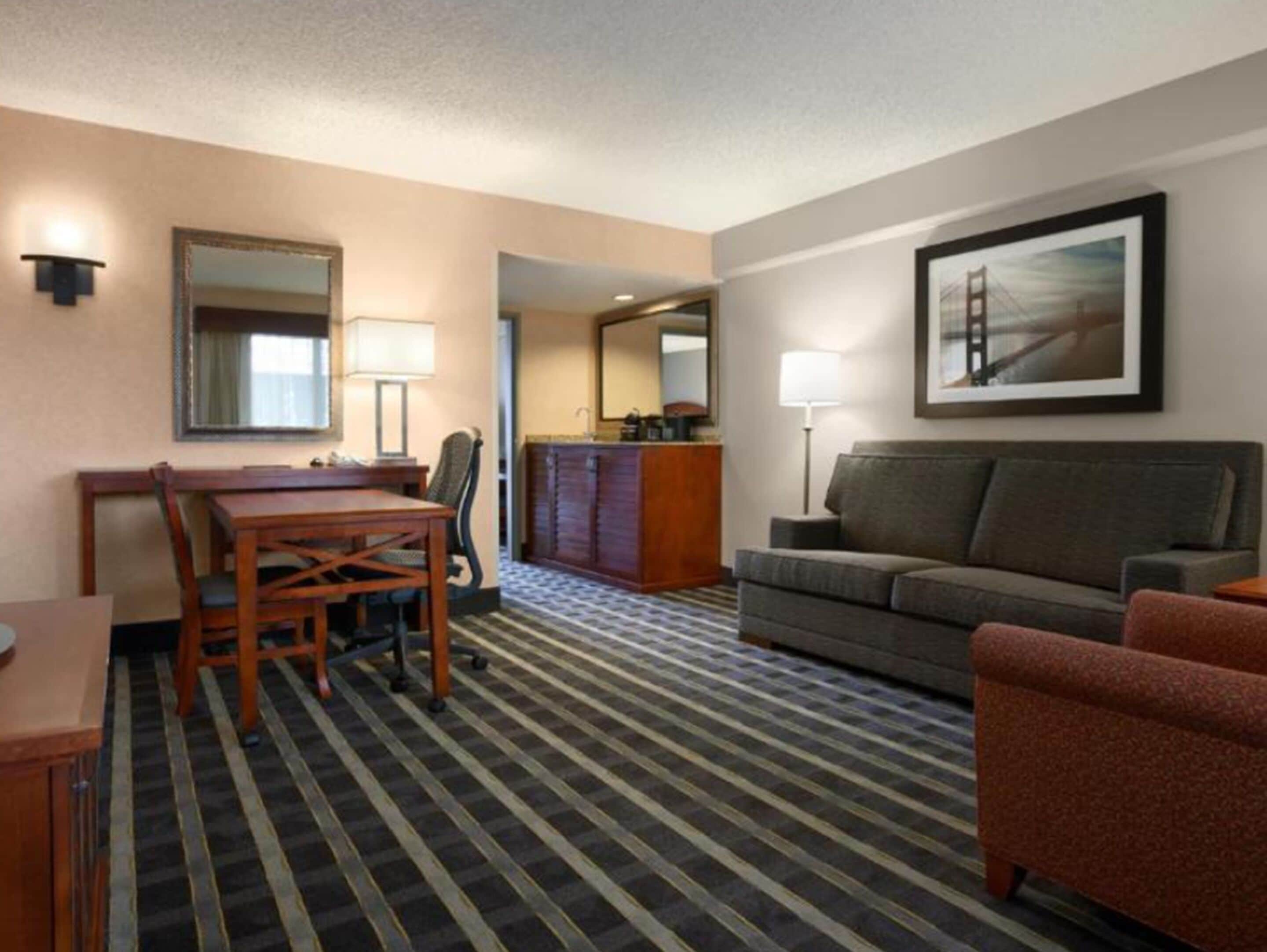 Ein Hotelzimmer mit Esstisch, Stühlen, einer Couch, einem kleinen Sessel, einer Lampe, einem Spiegel, einem gerahmten Bild und einem gemusterten Teppich.
