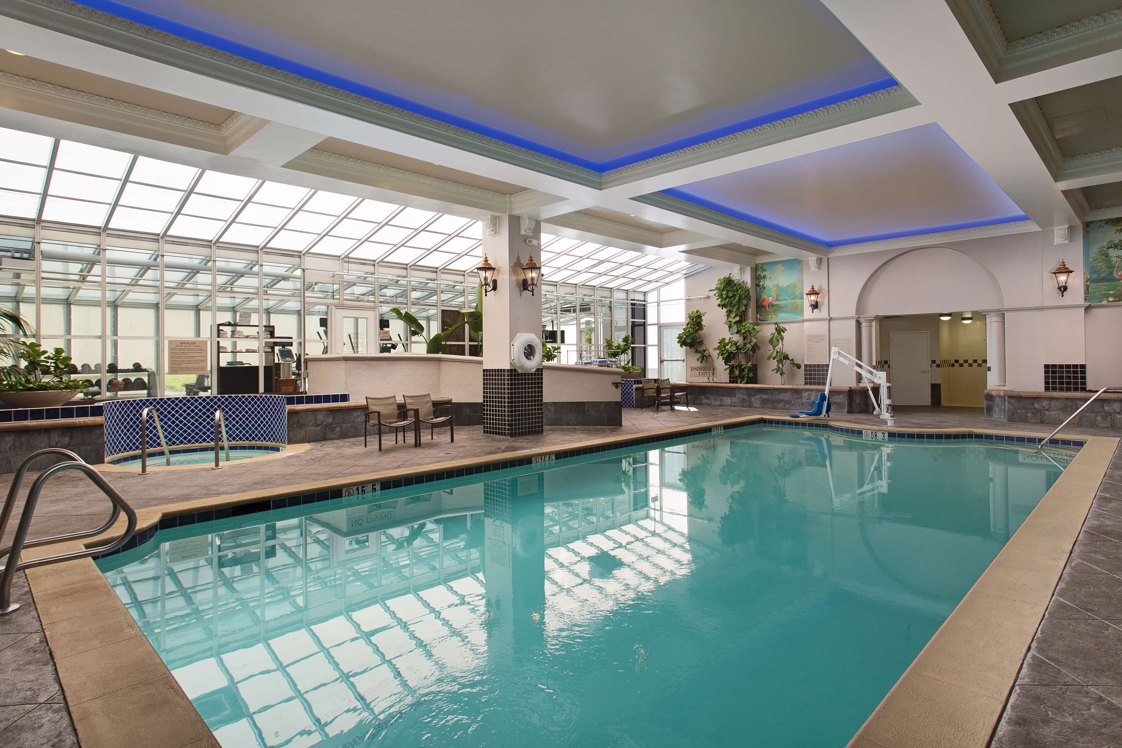 An indoor swimming pool with clear glass walls, a tiled deck, and a blue-lit ceiling.