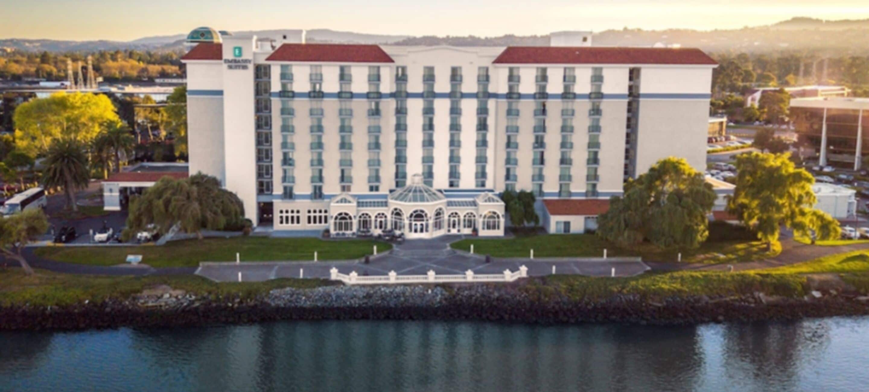 A large hotel with a central courtyard, a water body in front, and a view of the surrounding area.