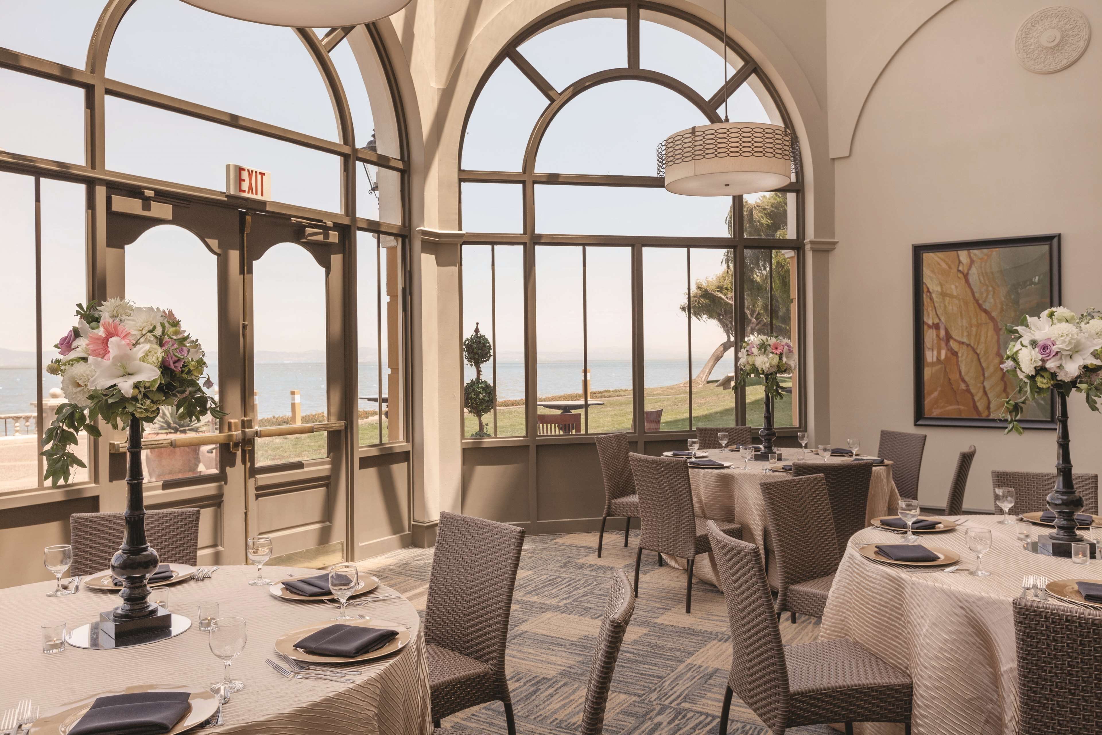 A restaurant with tables set for dining, large windows with an ocean view, and wicker chairs.