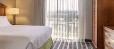 A hotel room with a bed, a nightstand, a lamp, a wooden wardrobe, and a large window with curtains.