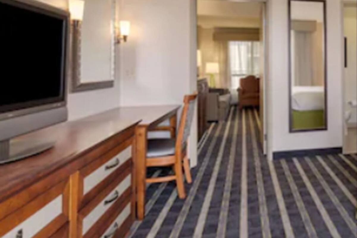 A hotel room with a television on a wooden dresser, a desk with chairs, a striped carpet, and a mirror on the wall.